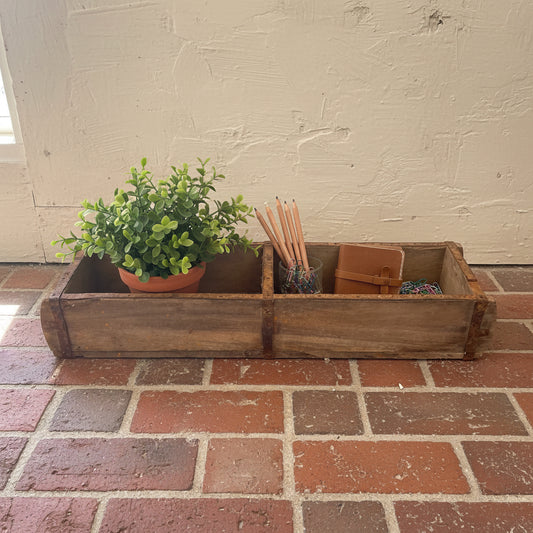 A double wooden Indian brickmould on a brick floor with a plant and pencils in the sections.