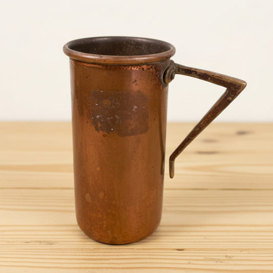 Vintage copper measuring cup on pine table against a white wall.
